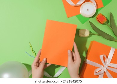 Both hands are holding an orange card, next to it are tulip branches, a gift box, ribbon and gift wrap on a pastel green background. Ideal space for letter design. - Powered by Shutterstock