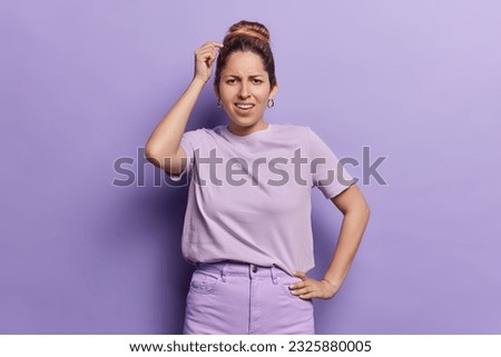 Bothered puzzled woman with combed hair scratches head stands indoor has dissatisfied expression tries to solve problem dressed in casual t shirt and trousers isolated over purple background