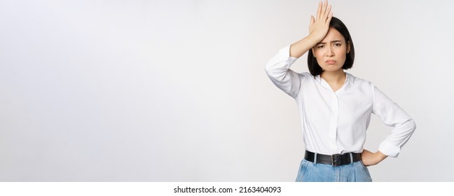 Bothered and tired asian woman, looking complicated, slap forehead, facepalm sign and grimacing upset, looking distressed at camera, white background - Shutterstock ID 2163404093