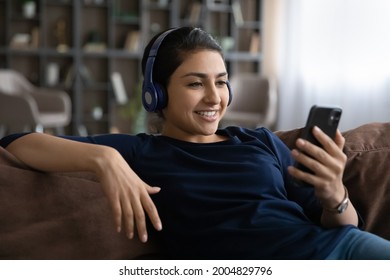 Bothering No One. Relaxed Indian Woman In Modern Earphones Sit On Comfy Couch Alone Watch Video Webinar Movie Online On Cellphone Screen. Carefree Young Lady Wearing Headset Listen To Music From Cell