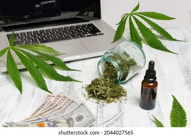 A Bottle Of Cannabis Oil, A Bottle With Dried Herb Splashing Around And Dollar Bills In Front Of A Laptop With Marijuana Leaves On White Background