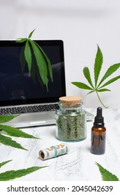 A Bottle Of Cannabis Oil, A Bottle Of Dried Herb And A Roll Of Dollar Bills In Front Of A Laptop With Marijuana Leaves On White Background