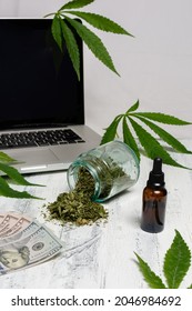 A Bottle Of Cannabis Oil, A Bottle With Dried Herb Splashing Around And Dollar Bills In Front Of A Laptop With Marijuana Leaves On White Background