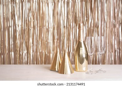 A Bottle Of Champagne, Glasses And Party Hats Are On The Table Against The Backdrop Of A Golden Tinsel Curtain.