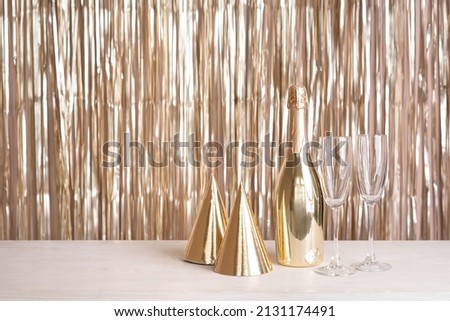 A bottle of champagne, glasses and party hats are on the table against the backdrop of a golden tinsel curtain.