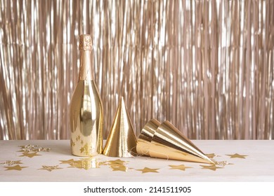 A Bottle Of Champagne And Party Hats Are On The Table Against The Backdrop Of A Golden Tinsel Curtain.