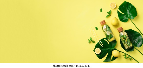 Bottle of detox water with mint, lemon and tropical monstera leaves on yellow background. Flat lay. Banner. Citrus lemonade. Summer fruit infused water. Top view with copy space