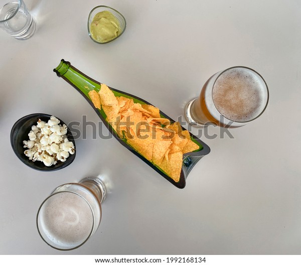 A bottle\
divided in half with tacos on a white table with beer, popcorn,\
guacamole sauce to dunk the tacos and a glass of water to ready\
everything as an aperitif before\
eating