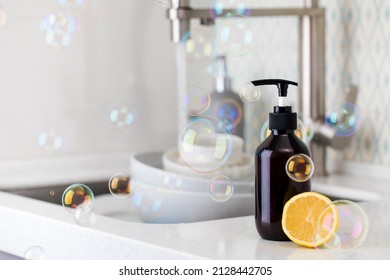 Bottle With Eco Friendly Dishwashing Detergent With Lemon On The Background Of A Sink With Dishes And Flying Bubbles.