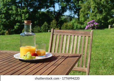 Bottle And Glass Of Orange Juice On Wooden Table With Chair. Blurred Green Trees And Hill Tops Background. Outdoor Breakfast With Fruit In Countryside. Picnic Or Garden Party Snack. No People.