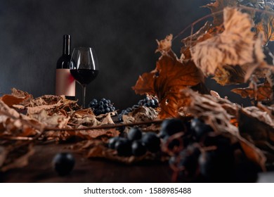 Bottle And Glass Of Red Wine On A Table With Dried Vine Leaves And Blue Grapes. On A Bottle Empty Old Paper Label, Free Space For Your Text. Selective Focus.