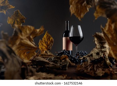 Bottle And Glass Of Red Wine On A Table With Dried Vine Leaves And Blue Grapes. On A Bottle Empty Old Paper Label. Selective Focus.