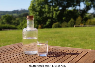 Bottle And Glass Of Water On Wooden Table With Chair. Blurred Green Trees And Hill Tops Background. Outdoor Breakfast In Countryside. Picnic, Garden Party Snack, Relaxation Concept. No People.