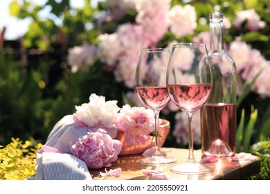 Bottle And Glasses Of Rose Wine Near Beautiful Peonies On Wooden Table In Garden