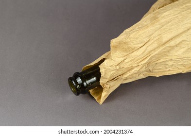 Bottle In Paper Bag Lying On Gray Background. Dark Empty Bottle Of Alcohol In A Crumpled Brown Bag. Close-up. Selective Focus.