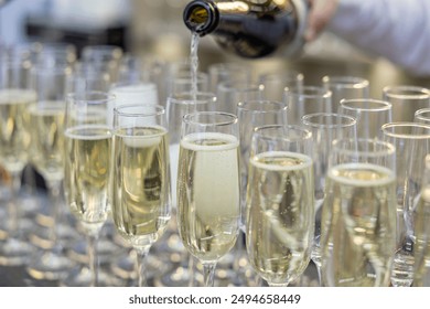 A bottle pours liquid into champagne glasses arranged in a row, suggesting a celebratory event - Powered by Shutterstock