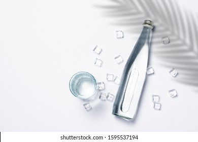 Bottle of pure crystal water with ice cubes on a white background under palm leaves and sunlight. Banner. Concept of thirst, heat, summer, tropic. Flat lay, top view