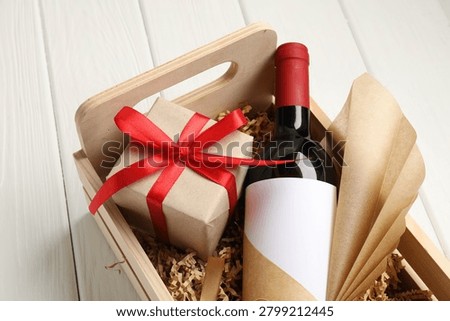 Bottle of red wine and gift box in crate on white wooden table, closeup