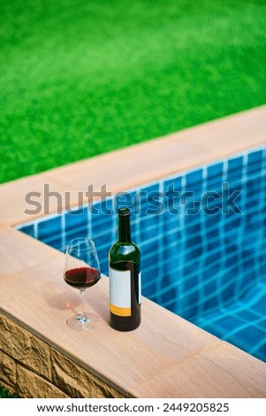 A bottle of red wine and a glass of wine are set on the edge of a clear blue pool, surrounded by a lush green lawn. The afternoon sun shines down, creating a relaxing and inviting atmosphere.