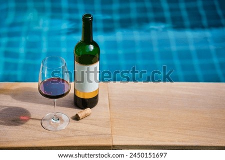 A bottle of red wine and a glass of wine are set on the edge of a clear blue pool, surrounded by a lush green lawn. The afternoon sun shines down, creating a relaxing and inviting atmosphere.
