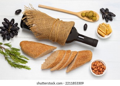 Bottle of red wine, green olives on wooden spoon. Crackers and nuts in bowls. Sliced baguette and sprig of grapes. Flat lay. White background. - Powered by Shutterstock