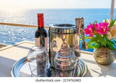 A Bottle Of Red Wine, An Ice Bucket And Two Stemless Glasses Next To A Bouquet Of Flowers On The Balcony Deck Of A Cruise Ship Cabin At Sea On A Summer Day.