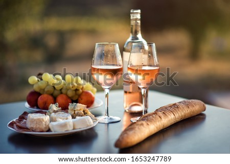 Bottle of rose wine and two full glasses of wine on table in heart of Provence, France with french bread, cheese, ham, grapes and peaches with olive trees on background in sunset. Travel in Provence