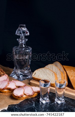 Bottle and shot glasses with vodka. Small snack of bread and meat near the shot glass. Slices of smoked meat or ham on brown packing paper. White wheat bread on wooden cutting board.