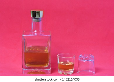 A Bottle Of Strong Alcohol. From Transparent Glass, A Square Form With A Golden Stopper. Near A Stack With A Drink Poured. The Second Stack Is Turned Upside Down. Background Of Coral Color.