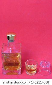 A Bottle Of Strong Alcohol. From Transparent Glass, A Square Form With A Golden Stopper. Near A Stack With A Drink Poured. The Second Stack Is Turned Upside Down. Background Of Coral Color.