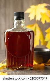 Bottle Of Tasty Maple Syrup, Bowl And Dry Leaves On Light Grey Table