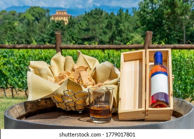A bottle of vin santo with its wooden box and a tray of cantuccini resting on top of a wooden barrel with the Tuscan countryside in the background, Pisa, Italy
