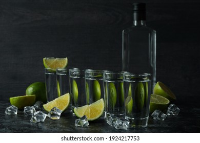 Bottle of vodka, shots, ice cubes and lime slices on black smokey table