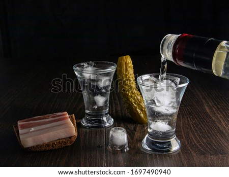A bottle of vodka, two misted glasses of cold vodka with ice on a wooden board with bacon and pickles.