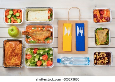 Bottle Of Water, Forks And Knives, A Paper Bag, Fish, Tomato And Feta Salad, A Pie, A Vegetable Roll, Baby Carrots, An Apple, Fruits, A Sandwich, Nuts And Dates On The Wooden Surface. Takeout Food.