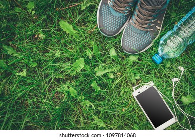 Bottle of water, mobile phone with headphones and sneakers on grass background.