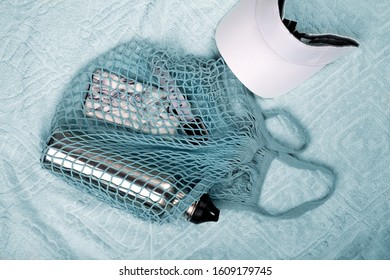 Bottle Of Water, Mobile Phone And Smart Watch In Blue String Mesh Reusable Bag And White Visor On The Beach Towel