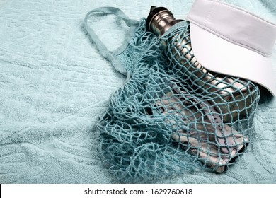 Bottle Of Water, Mobile Phone And Smart Watch In Blue String Mesh Reusable Bag And White Visor On The Beach Towel