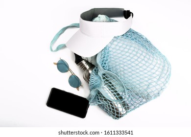 Bottle Of Water, Mobile Phone, Sun Glasses, Beach Towel And White Visor In Blue String Mesh Reusable Bag On White Background