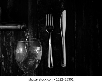 A Bottle Of Wine Pouring Into A Glass With Ice Cubes, On A Blurred Overhead Knife And Fork Background.