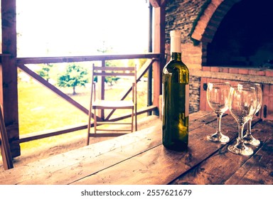 A bottle of wine and three empty wine glasses on a rustic wooden table in an outdoor patio setting. Warm sunlight and a countryside backdrop create a cozy and inviting atmosphere. - Powered by Shutterstock