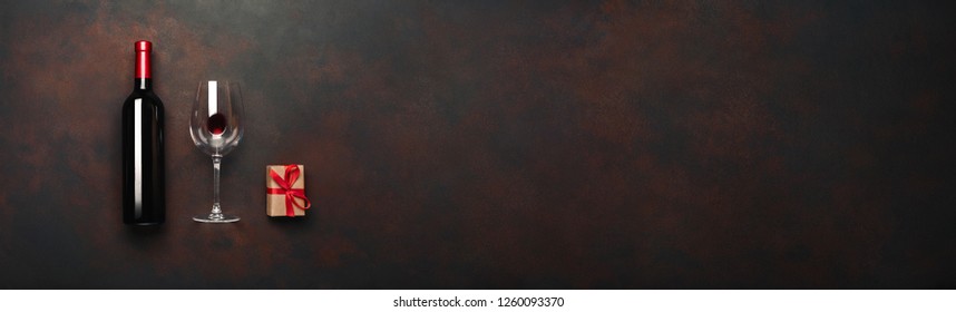 Bottle of wine with wineglass and gift box on rusty brown background. Panoramic top view with copy space for your text.