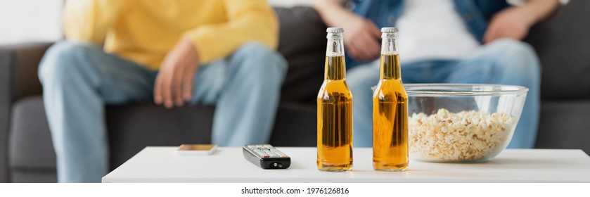 bottles of beer, smartphone, tv remote and popcorn near friends sitting on blurred background, banner - Powered by Shutterstock