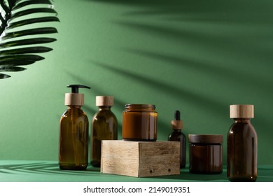 Bottles for cosmetics and cream on a pedestal, green background with leaves and shadow. Brown glass jars with wooden lids. Eco concept. Container for shampoo, oil. Bottle with dispenser and pipette.