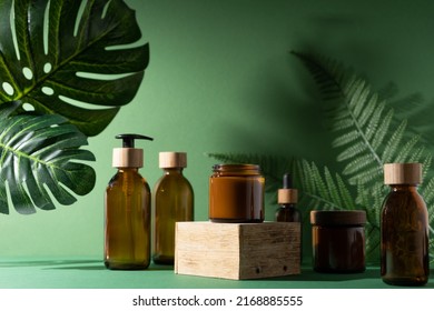 Bottles for cosmetics and cream on a pedestal, green background with leaves and shadow. Brown glass jars with wooden lids. Eco concept. Container for shampoo, oil. Bottle with dispenser and pipette.