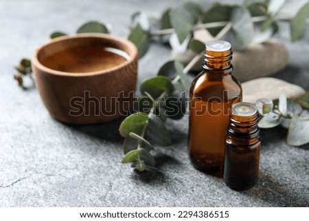 Bottles of eucalyptus essential oil, wooden bowl and plant branches on light grey table, space for text