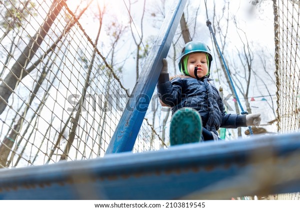 Bottom POV view brave courage little toddler child\
boy wear safety equipment helmet enjoy passing obstacle course\
forest rope adventure park on cold winter day. Active outside\
leisure amusement camp