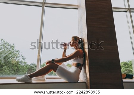 Bottom side view full body size young sporty woman 20s wear white sportswear earphones listen music drink bottle water sit on window windowsill of gym near treadmill rest indoors People sport concept.