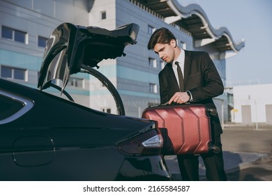 Bottom side view serious young traveler businessman young man in black suit stand outside at international airport terminal put suitcase into car trunk booking taxi Air flight business trip concept.