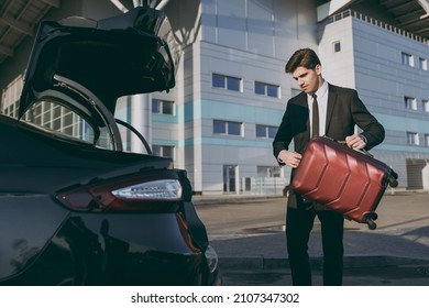 Bottom side view young traveler businessman young man in black dinner suit stand outside at international airport terminal put suitcase into car trunk booking taxi Air flight business trip concept.
